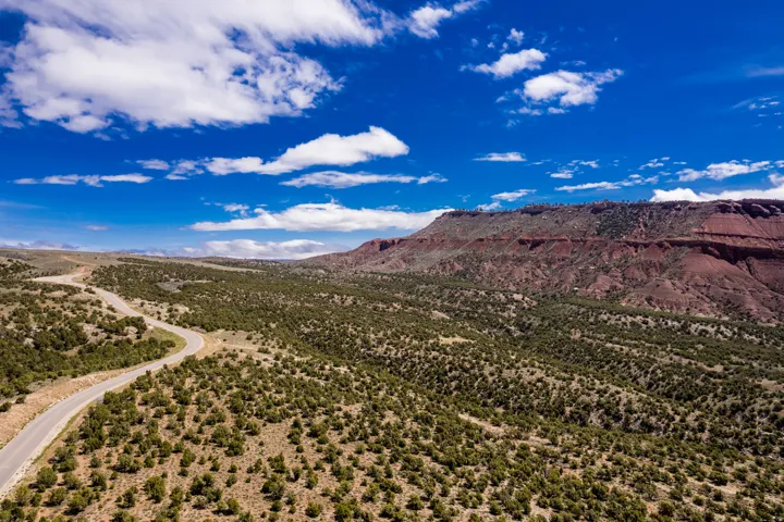 Sweeping landscape featuring a winding paved road, red rock formations, abundant low-lying green vegetation, and a clear blue sky with scattered clouds