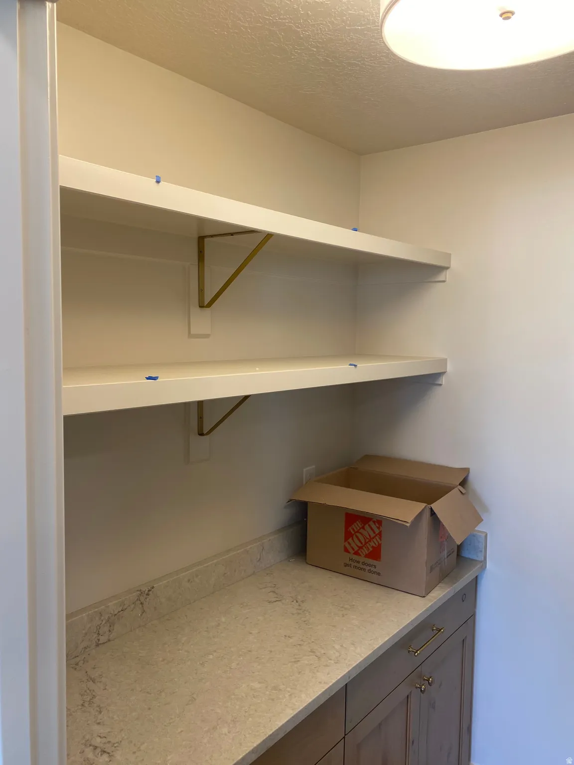 Organized pantry featuring two white floating shelves with brass-tone brackets, a light-toned countertop, and wood-finish lower cabinetry with brass hardware