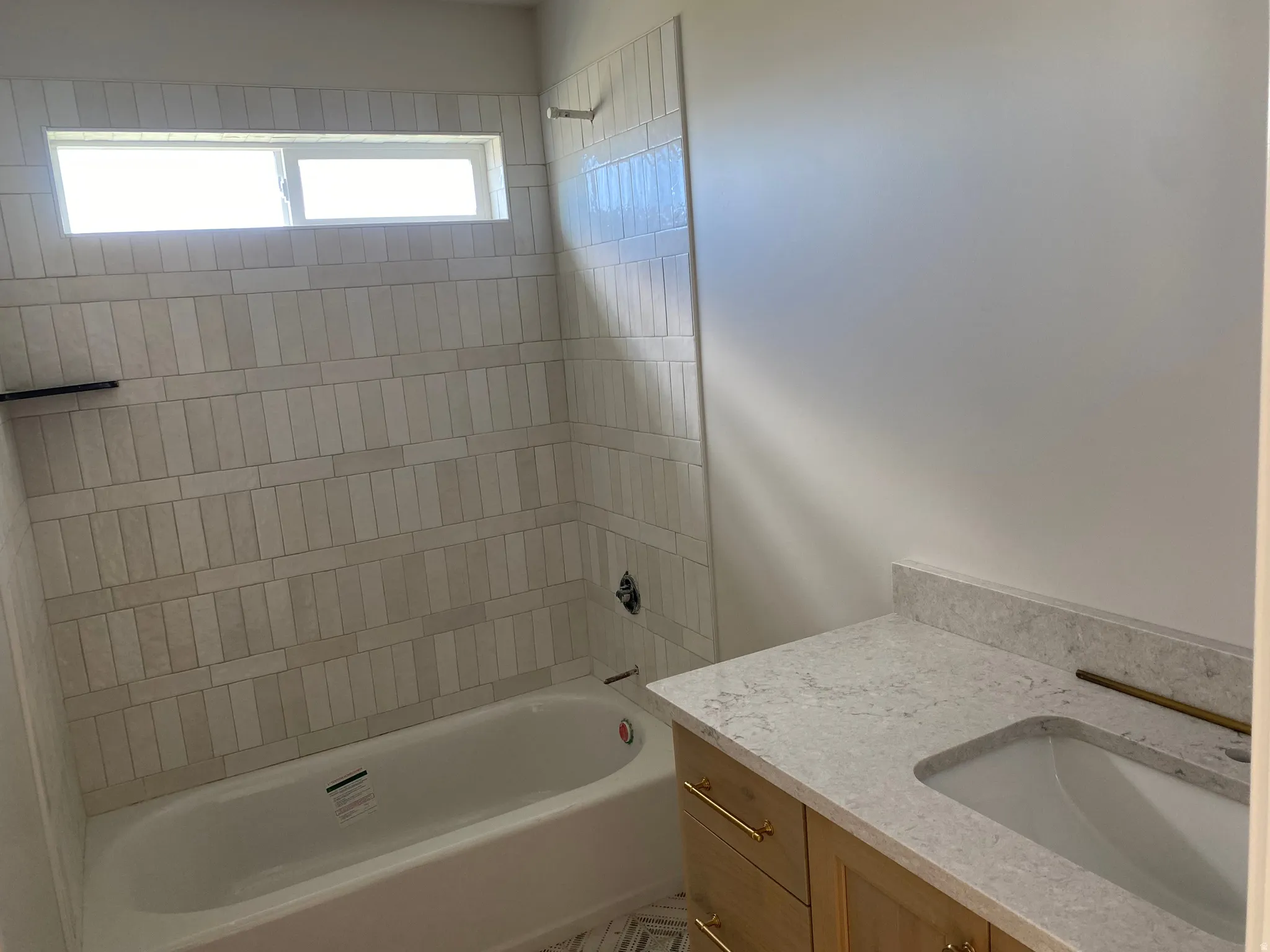 Bathroom featuring a built-in bathtub with subway tile surround, integrated window, light wood-finish vanity with gold-tone hardware, and a stone countertop with undermount sink