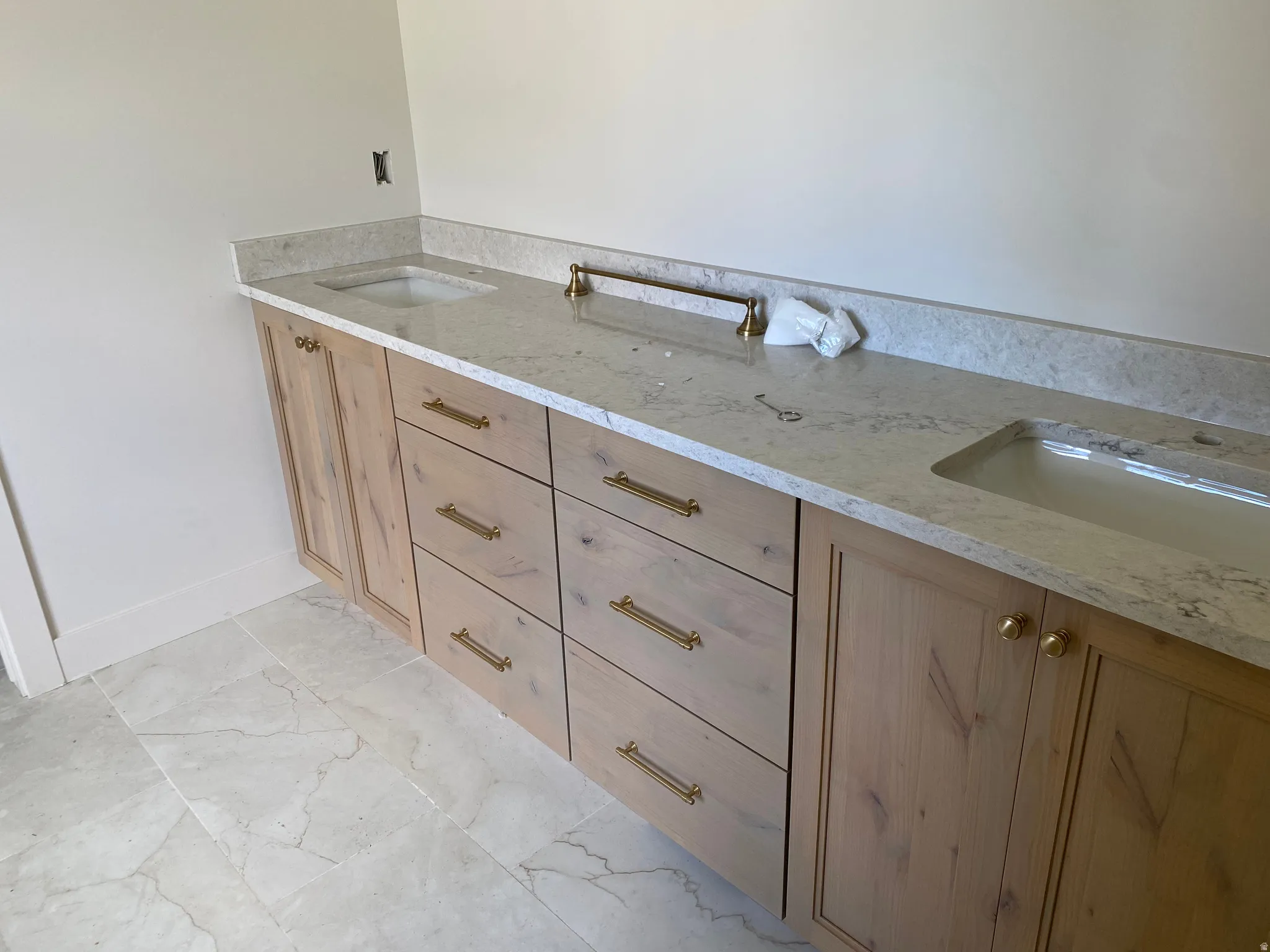 Double vanity featuring light wood cabinetry, gold-tone hardware, and light-toned stone countertops with integrated sinks