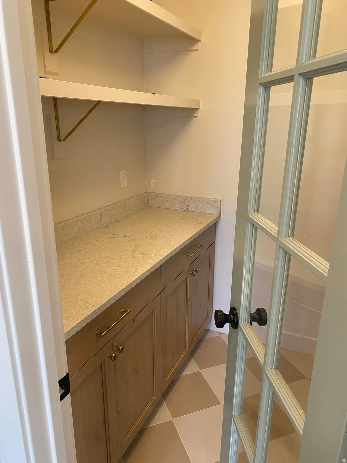 Pantry featuring checkerboard floor tiles, built-in lower cabinetry with gold-tone hardware, a light-toned countertop, and open shelving with gold-tone brackets