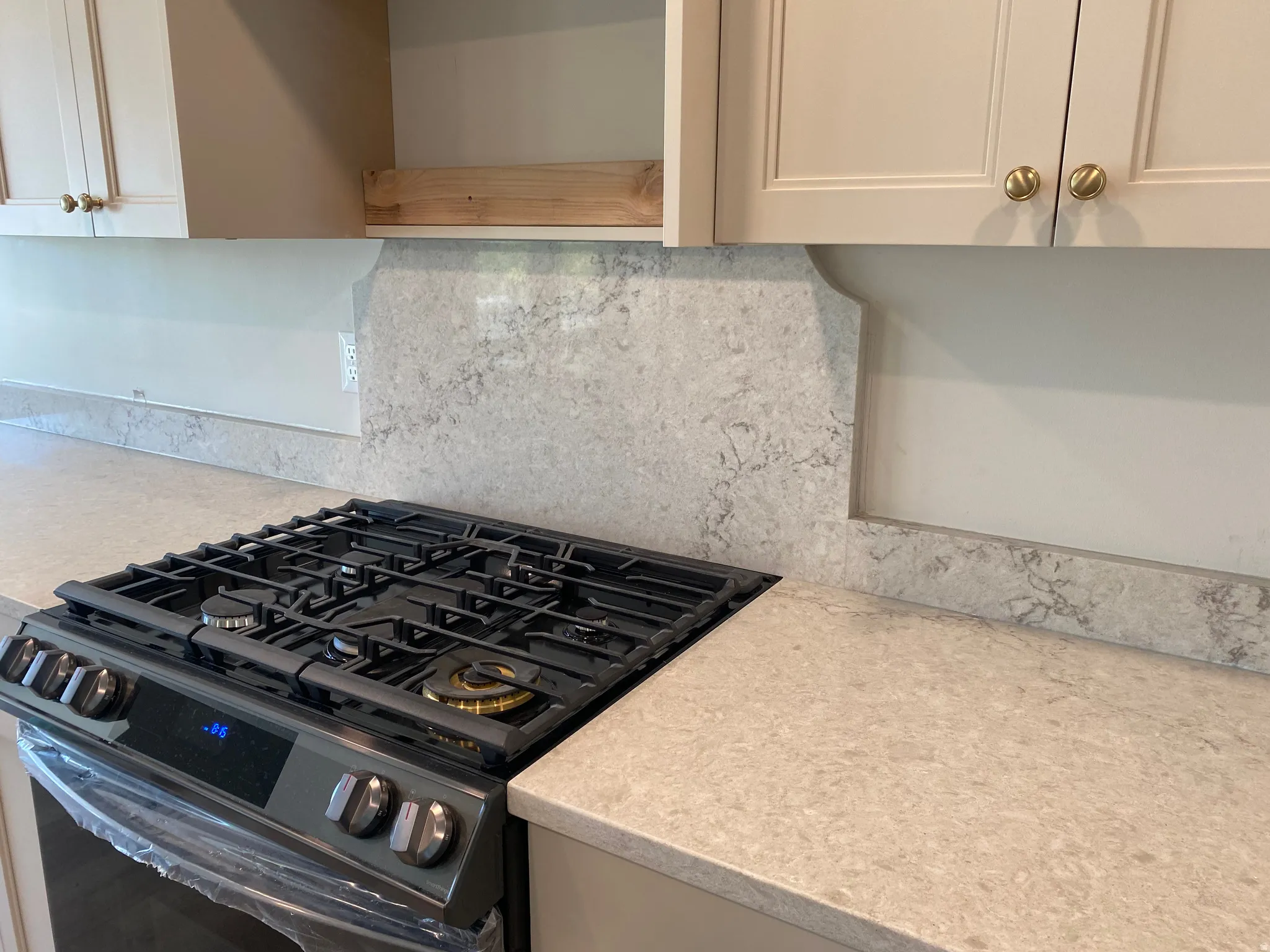 Built-in gas range with five burners, light-toned countertops, stone-finish backsplash, and neutral cabinetry with brass hardware
