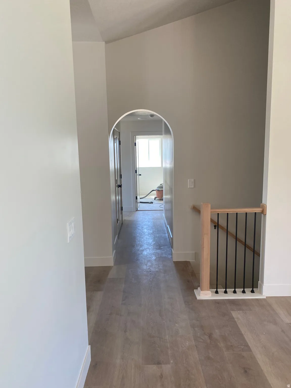 Arched doorway opening to a hallway featuring wood-finish flooring, crisp white baseboards, and a natural wood banister with black iron balusters