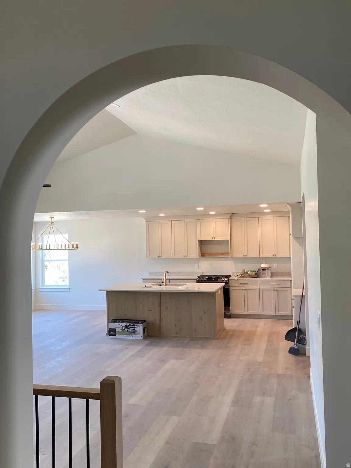 Arched entryway framing a kitchen with light wood-finish flooring, recessed lighting, a central island with a sink, and light-toned cabinetry
