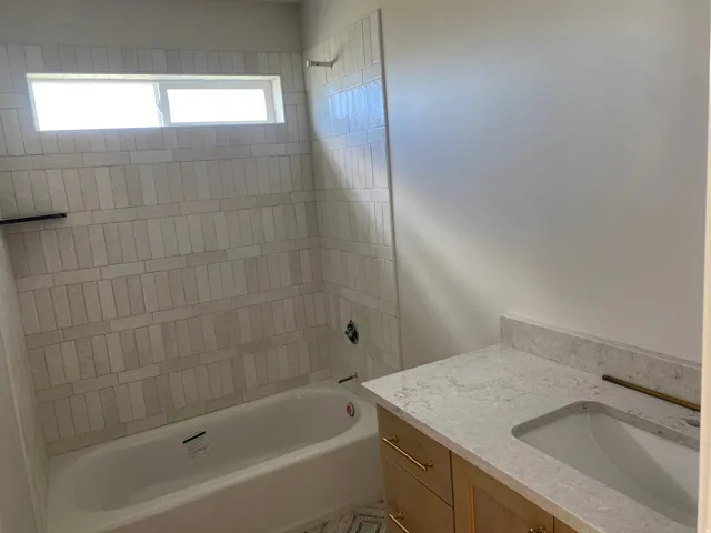 Bathroom featuring a built-in bathtub with subway tile surround, integrated window, light wood-finish vanity with gold-tone hardware, and a stone countertop with undermount sink