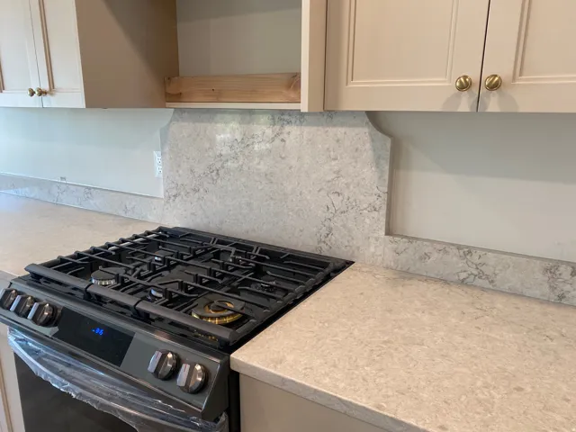Built-in gas range with five burners, light-toned countertops, stone-finish backsplash, and neutral cabinetry with brass hardware