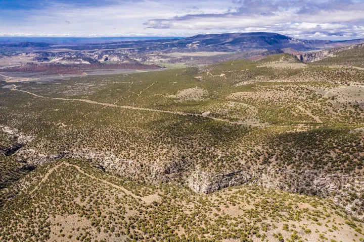 Expansive natural landscape featuring undulating terrain, sparse green vegetation, and distant mountain ranges under a partly cloudy sky