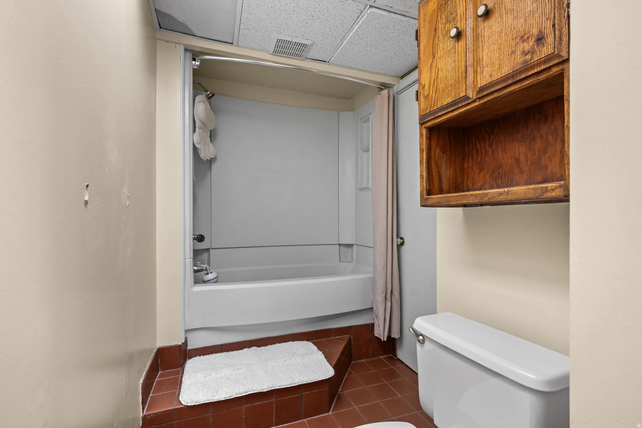 Bathroom with a white tub and shower combination, terracotta-toned tile flooring, and a drop ceiling
