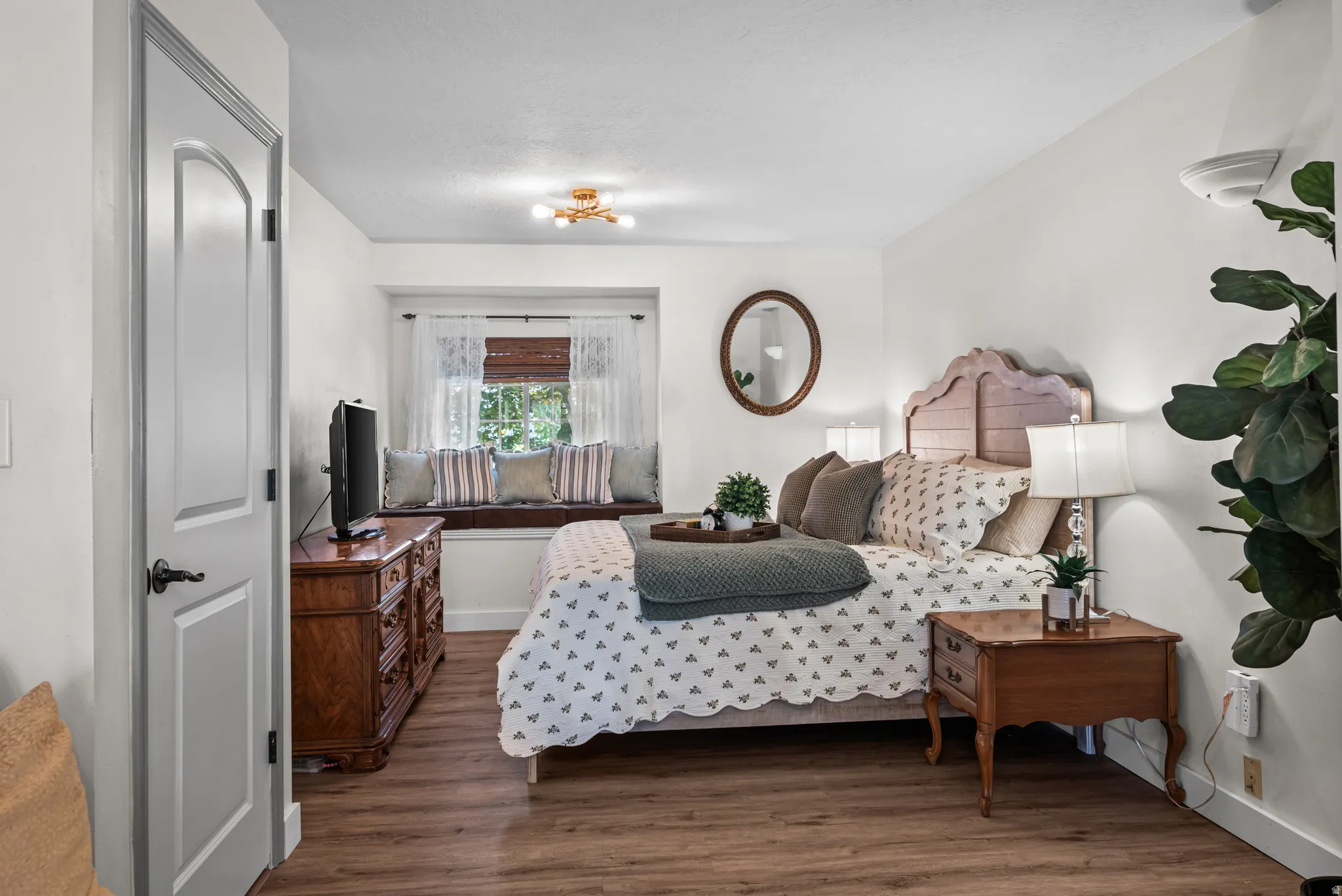 Bedroom featuring wood-finish flooring, a built-in window seat, a paneled door with dark hardware, a light fixture, and an oval mirror