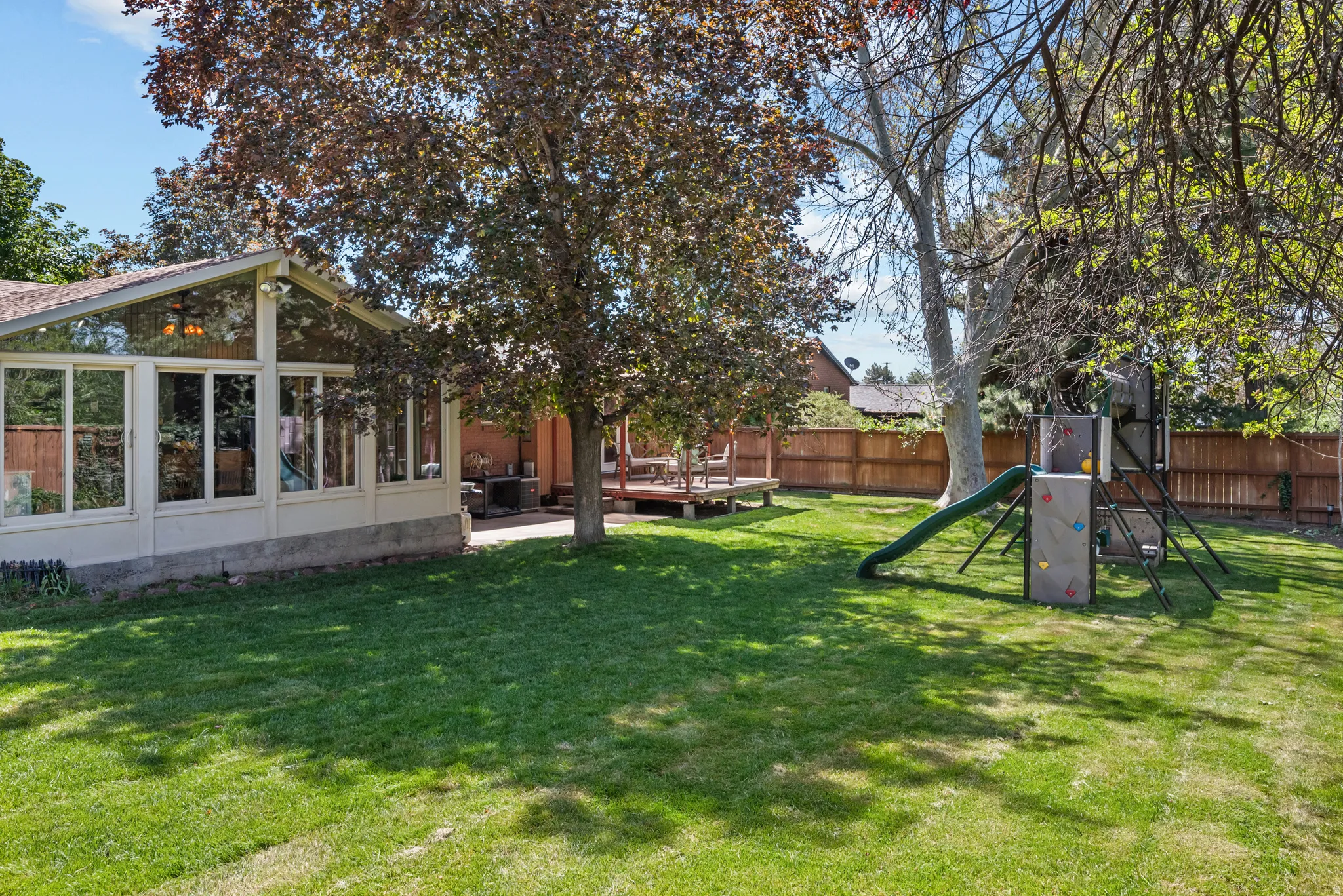 Expansive backyard featuring a manicured lawn, a sunroom with large windows, a raised wooden deck, and mature trees providing shade