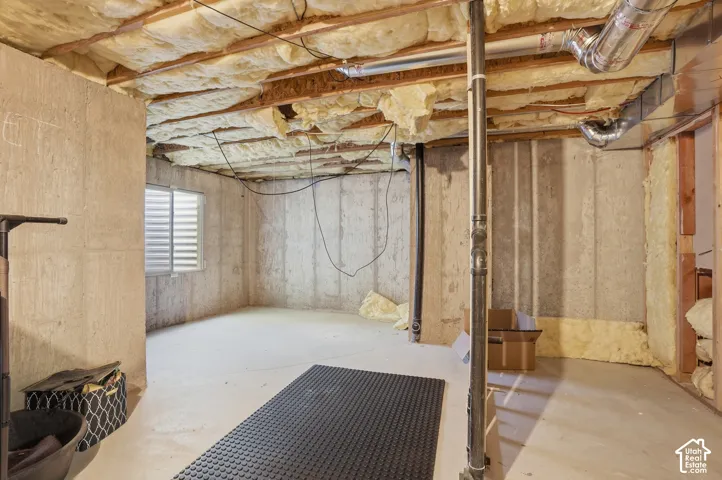 Unfinished basement featuring concrete walls and flooring, exposed ceiling joists with insulation, and a window with horizontal blinds. This could be a converted to a second owners suite. Room can be finished, rough plumbing is located in this room.