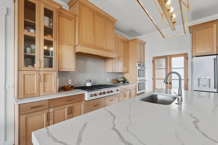 Spacious kitchen featuring a large island with an undermount sink, light wood cabinetry, white countertops, built-in double ovens, and a gas range with a herringbone tile backsplash