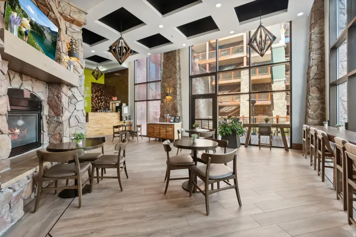 Open-concept common area featuring a stone fireplace, wood-finish flooring, full-height windows, a built-in bar, and coffered ceilings with geometric pendant lighting