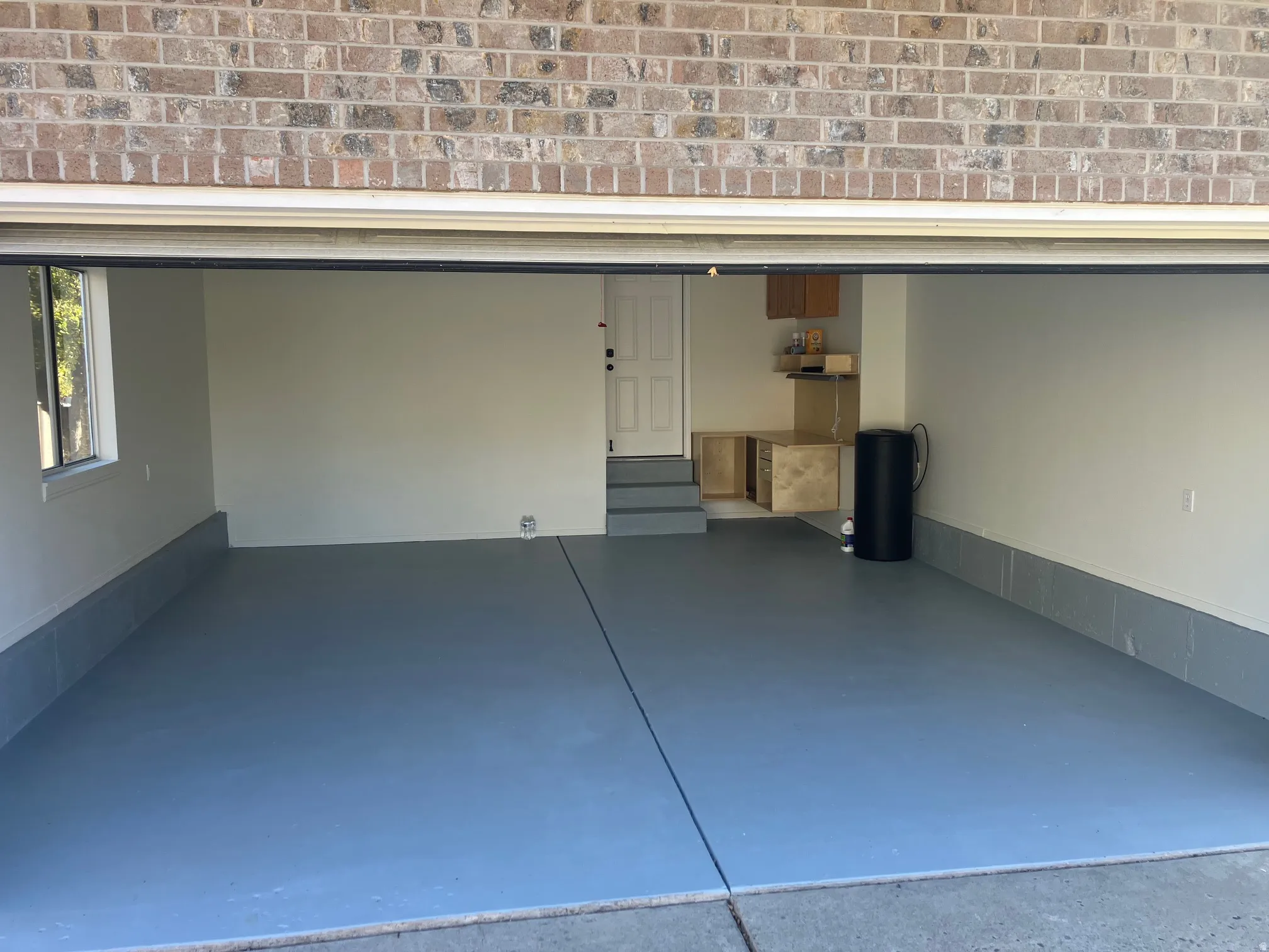 Spacious garage featuring a finished concrete floor, light-toned walls, a window, built-in cabinetry, and brick exterior detailing.  Storage shelf upon overhead garage door.