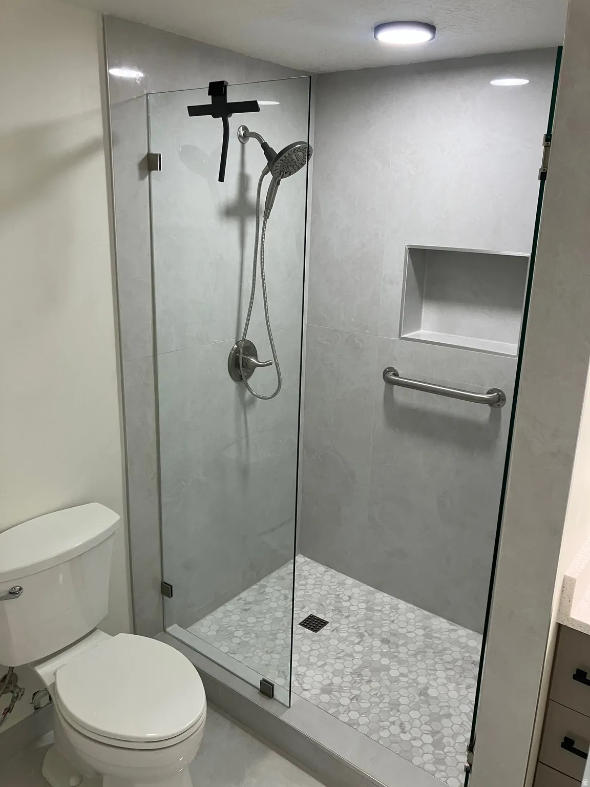 Frameless glass shower enclosure featuring large format wall tiles, hexagonal mosaic floor tiles, a built-in shower niche, and a brushed nickel shower fixture