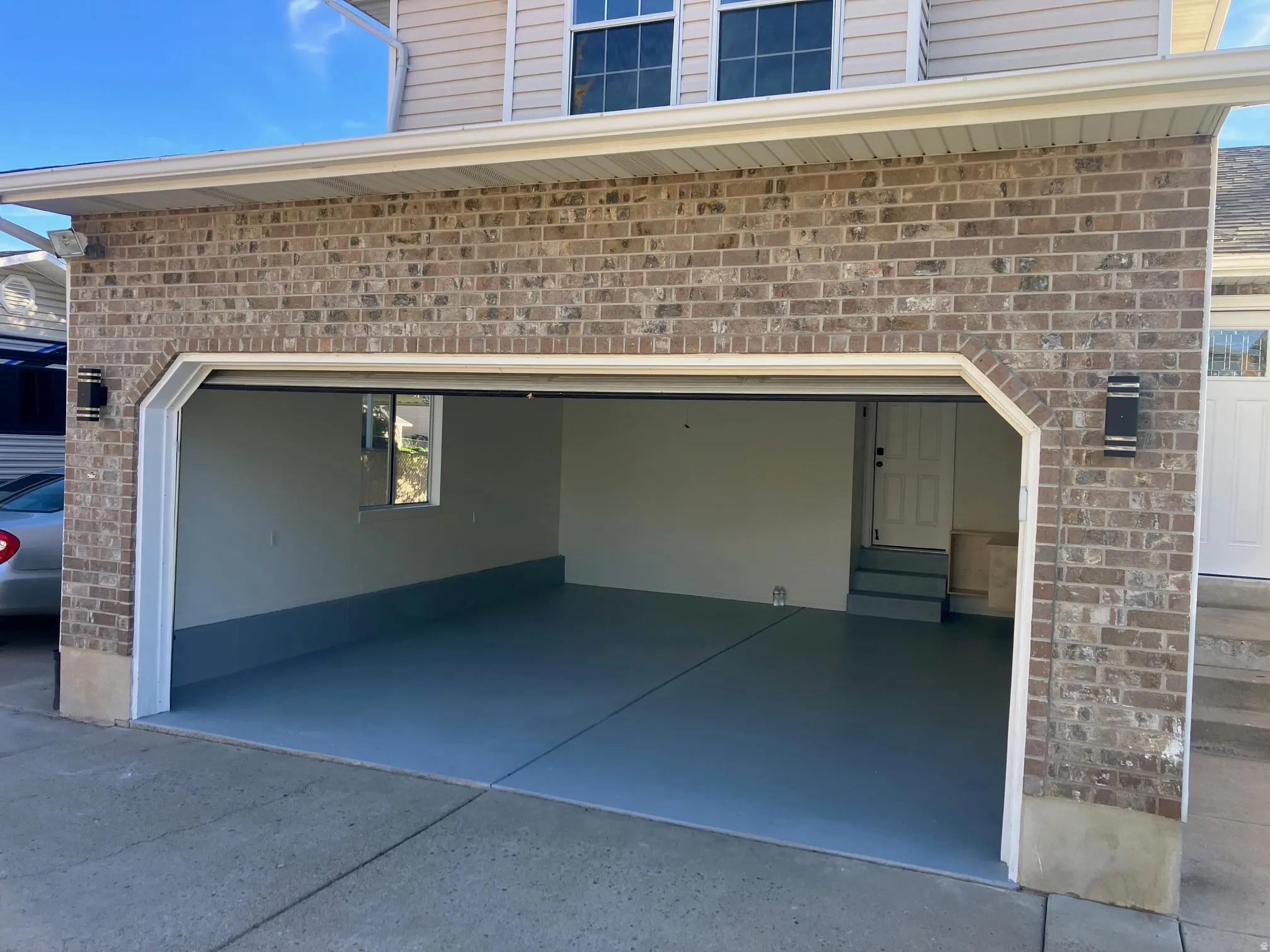 Two-car garage featuring brick exterior, painted concrete flooring, exterior wall sconces, and interior access door