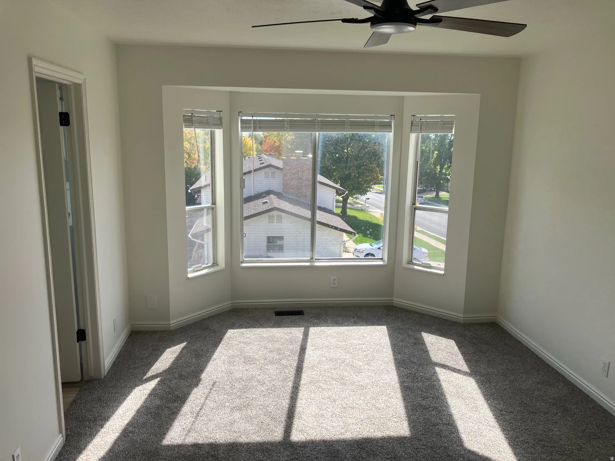 Light-filled room featuring a bay window, plush grey carpeting, white wall paint, a black ceiling fan, and white trim