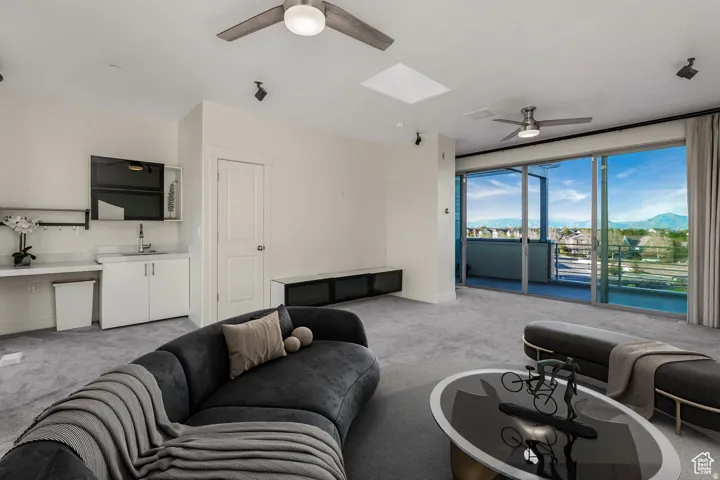 Spacious carpeted room featuring a wet bar with a single basin sink and white cabinetry, two ceiling fans, a skylight, and expansive glass sliding doors opening to a balcony