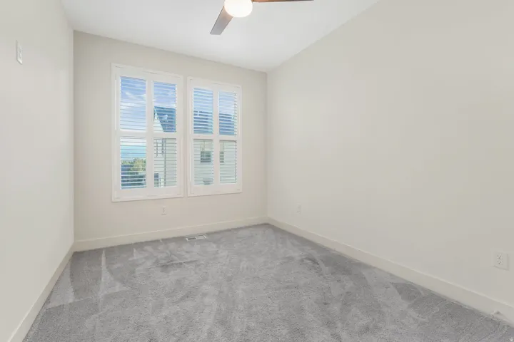 Neutral-toned room featuring a ceiling fan, dual windows with white plantation shutters, and gray patterned carpet
