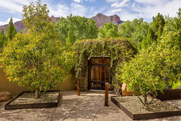 Enclosed courtyard featuring a rustic wooden gate, mature trees with metal-edged planters, gravel ground cover, and a stucco privacy wall