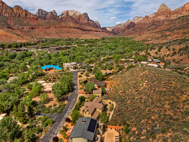 Expansive aerial capturing a neighborhood with varied architectural styles set against a backdrop of towering red rock formations and a lush green valley