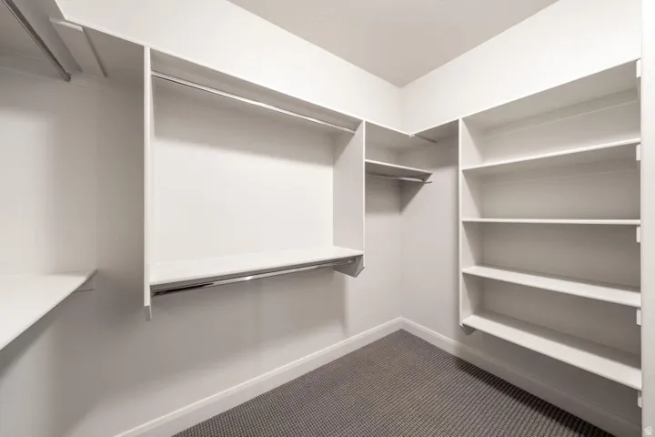 Walk-in closet featuring white built-in shelving, multiple hanging rods, and grey patterned carpeting