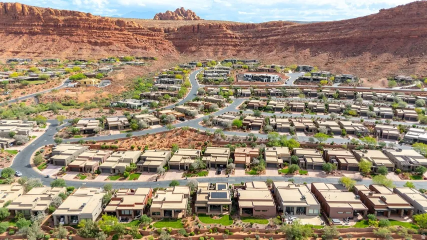 Expansive residential development featuring contemporary architecture, multiple single-story residences, paved streets, and varied landscaping, all set against a backdrop of natural red rock formations