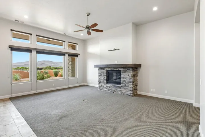 Spacious interior featuring a stacked stone fireplace with a substantial wood mantel, a ceiling fan, recessed lighting, and large windows with integrated shades offering expansive outdoor scenery
