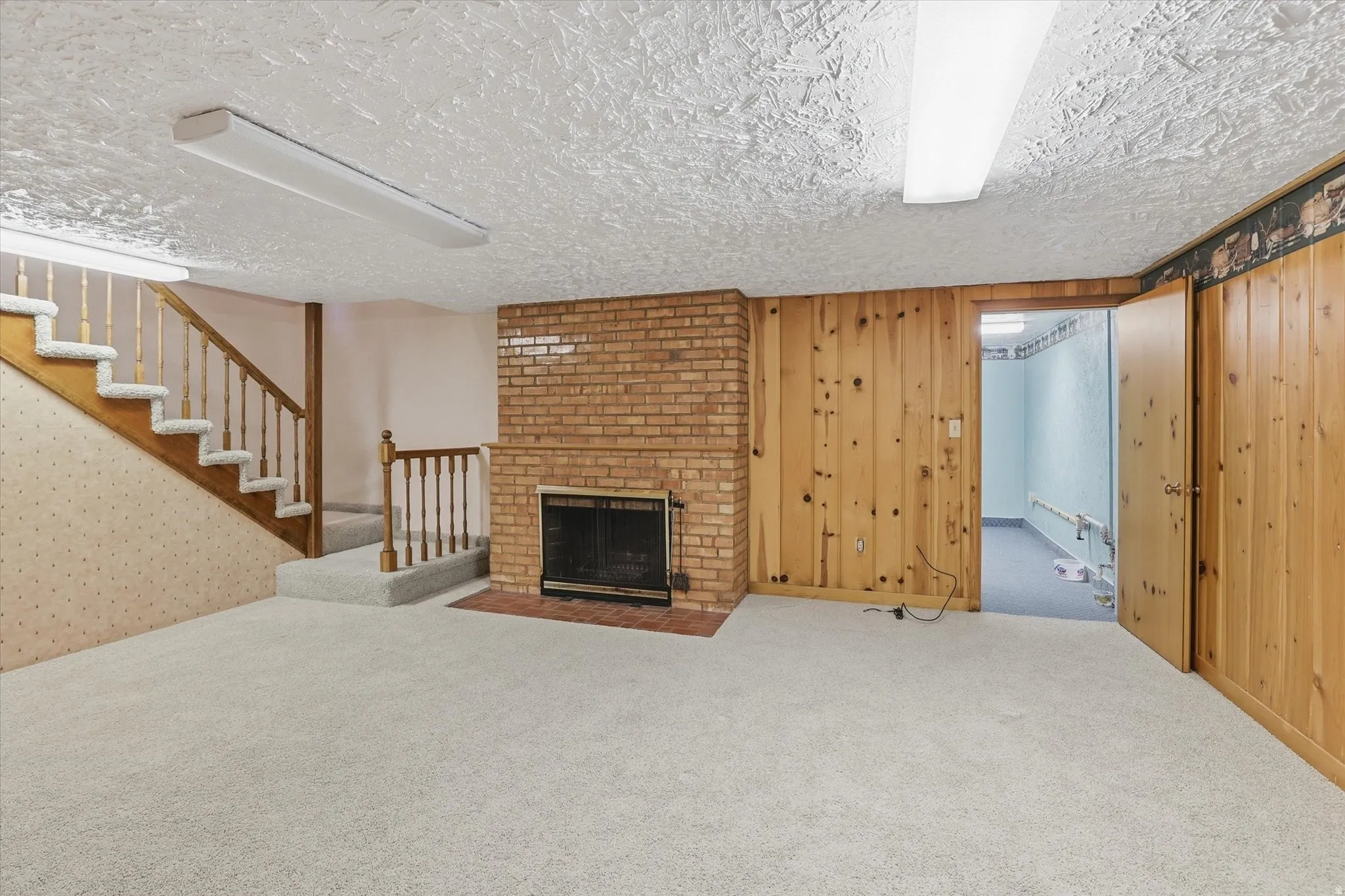Spacious lower-level room featuring a brick fireplace, light-colored carpet, wood-paneled walls, a staircase with wood railings, and fluorescent ceiling lights