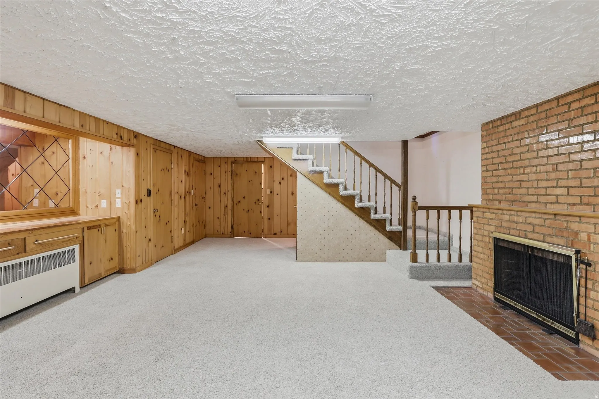 Spacious carpeted room featuring a brick fireplace with a metal screen, wood paneling with built-in cabinetry, and a carpeted staircase with wood railings