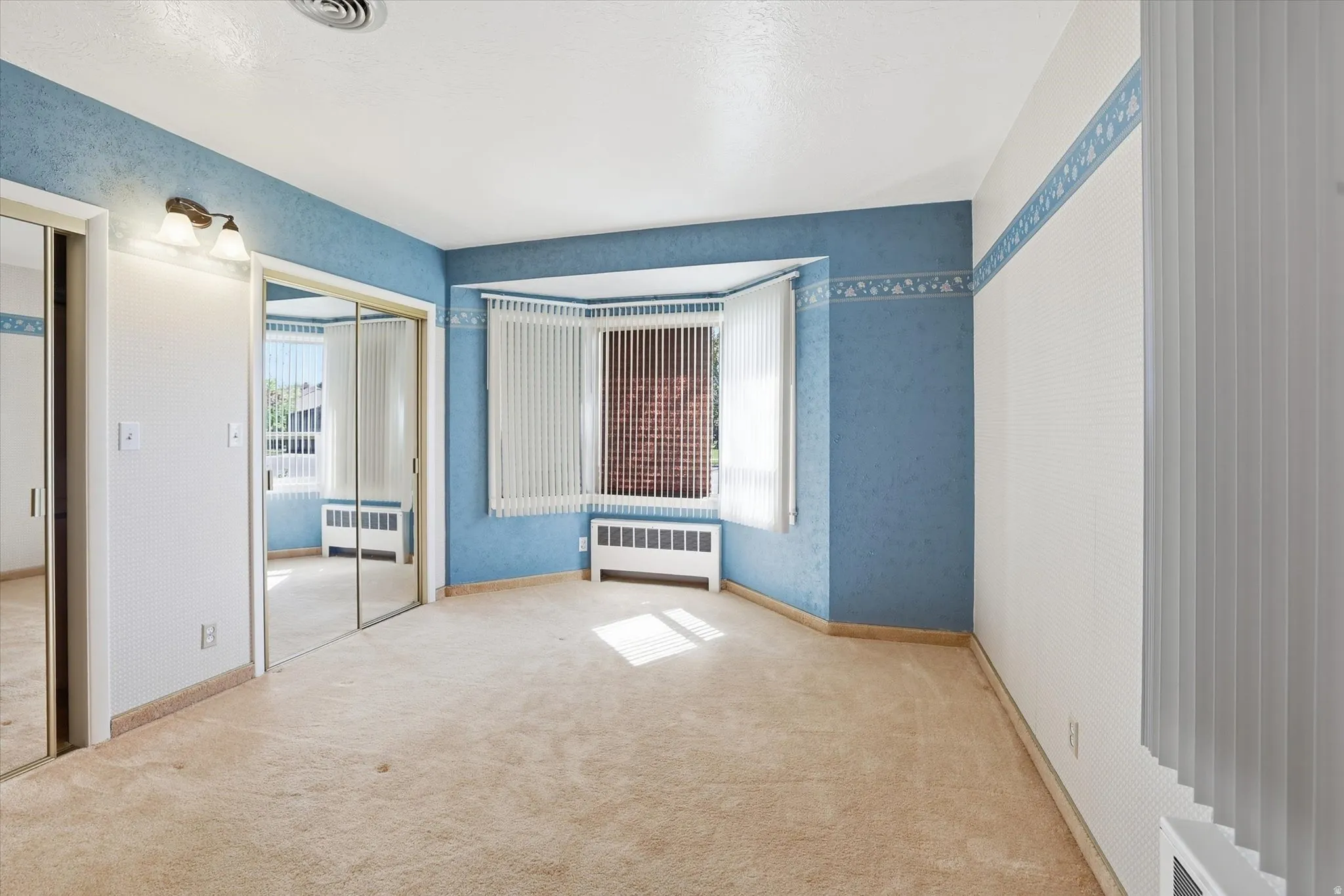 Carpeted room featuring a bay window with vertical blinds, wall-mounted lighting fixture, and mirrored closet doors