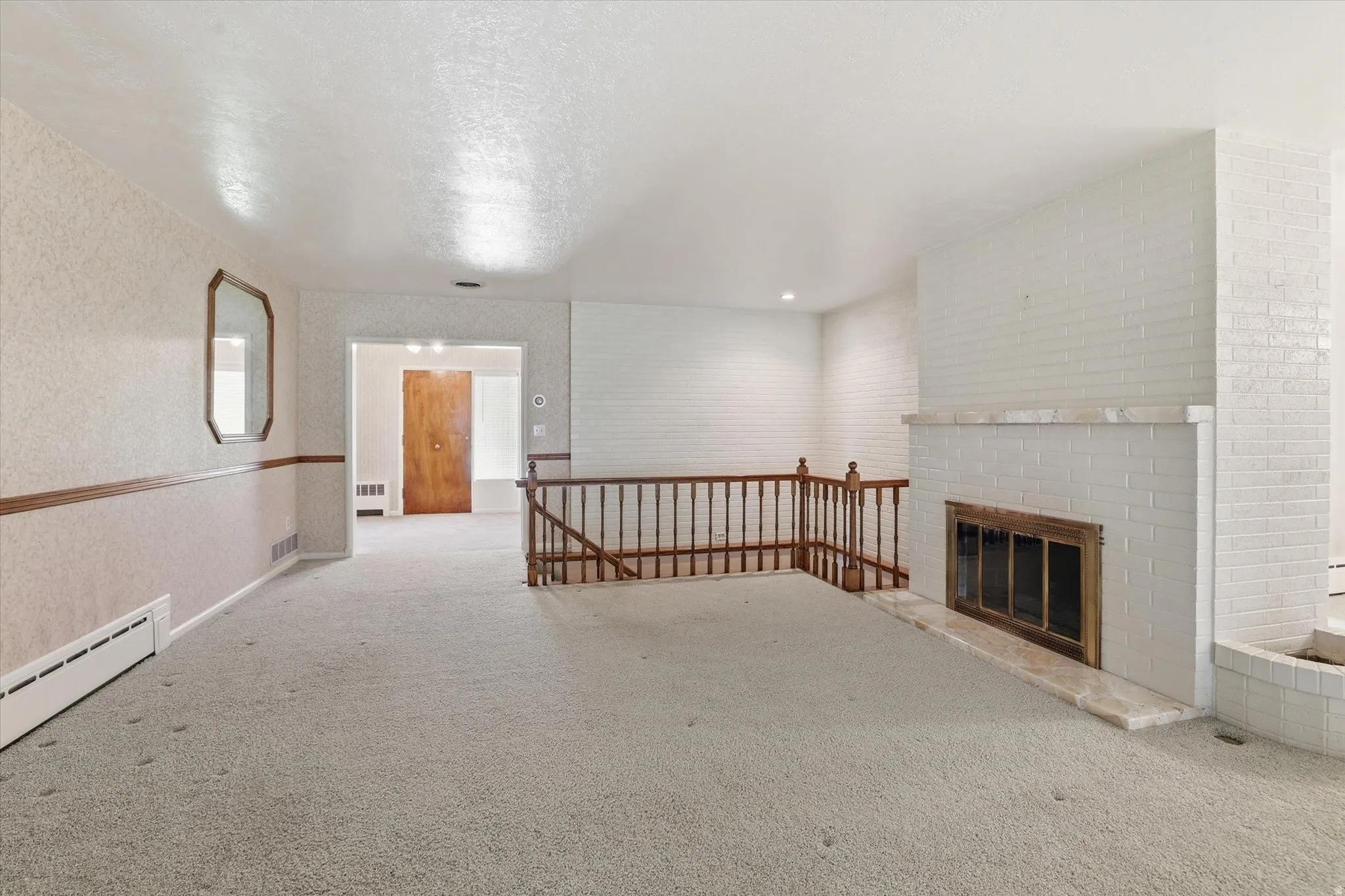 Living area featuring light-toned carpeting, white painted brick fireplace with a stone hearth, and natural wood railing