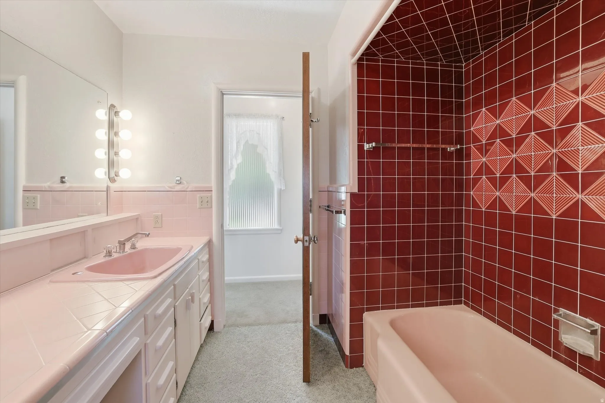 Bathroom featuring a pink vanity with a single basin sink, large mirror with vertical strip lighting, and a bathtub with red tiled walls and geometric accent patterns