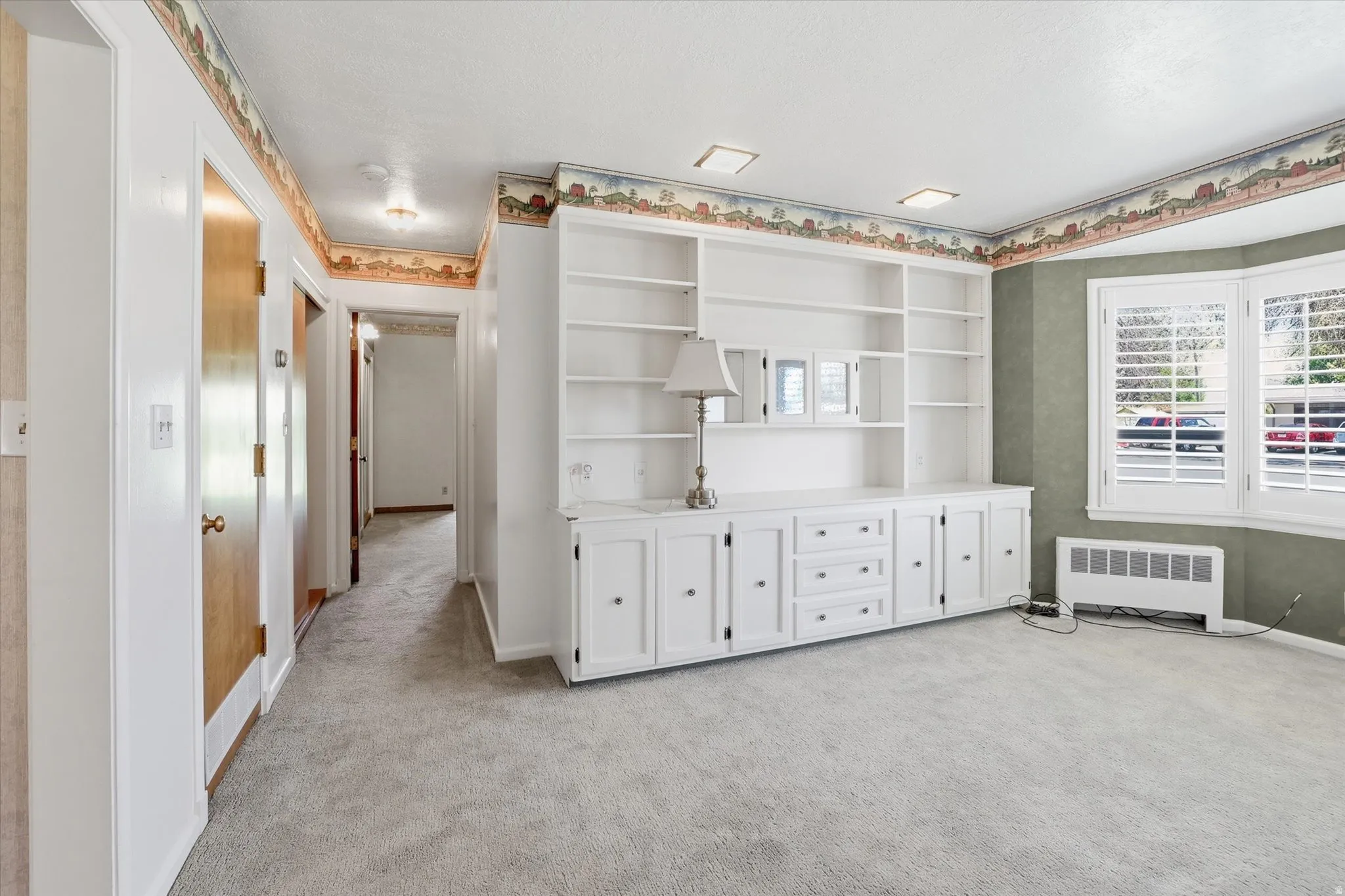 Integrated white cabinetry with shelving, a bay window featuring white plantation shutters, light gray carpet flooring, recessed ceiling lighting, and a corridor with natural wood doors