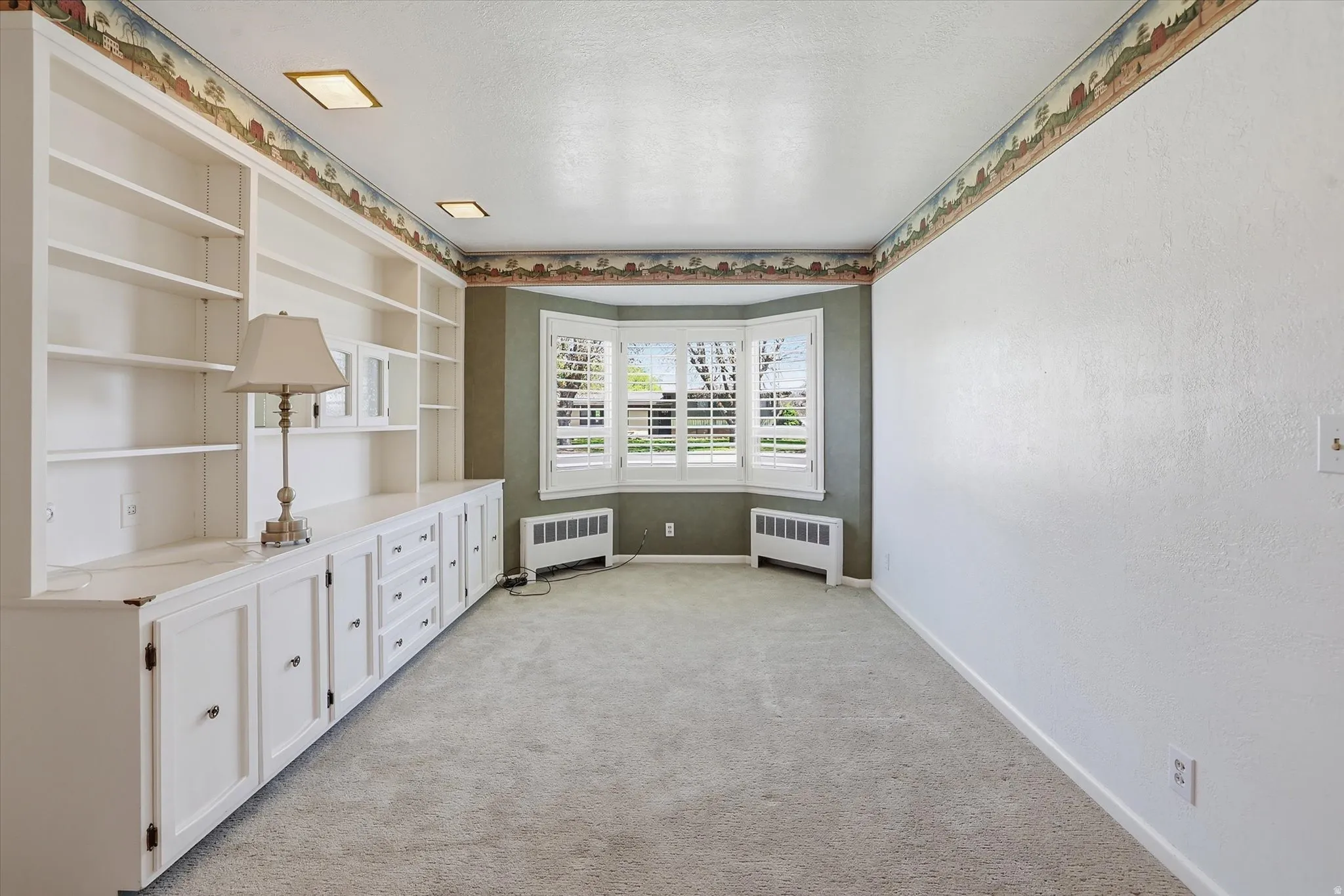 Room featuring a built-in white shelving unit with lower cabinetry, bay windows with plantation shutters, two baseboard heating units, and recessed ceiling lighting