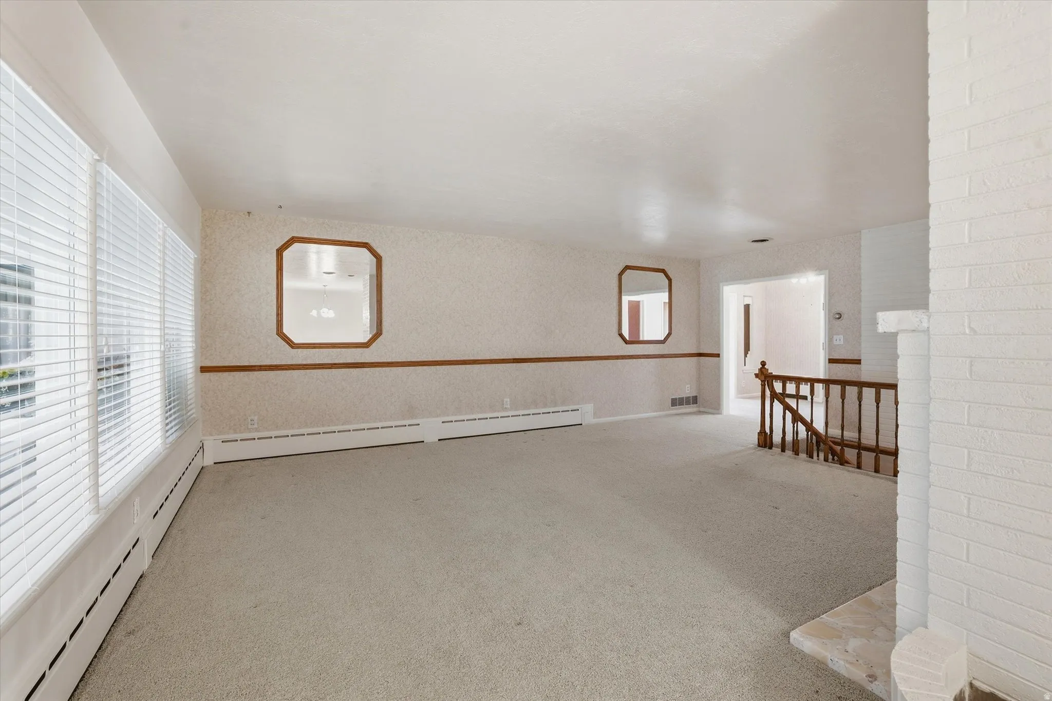 Expansive carpeted room featuring a full wall of windows with blinds, a white brick-finish fireplace, and wood-finish railing