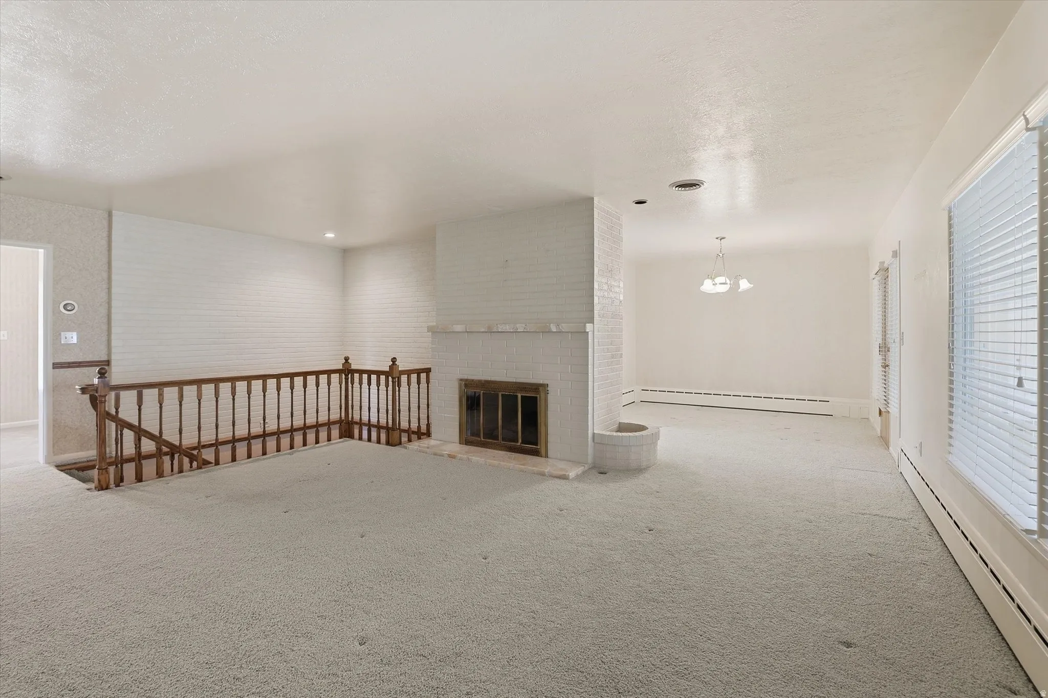 Spacious interior featuring light-colored carpeting, a white painted brick fireplace with hearth, and a wooden banister overlooking a lower level