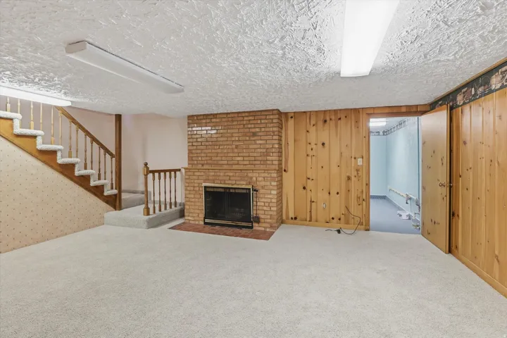 Spacious lower-level room featuring a brick fireplace, light-colored carpet, wood-paneled walls, a staircase with wood railings, and fluorescent ceiling lights
