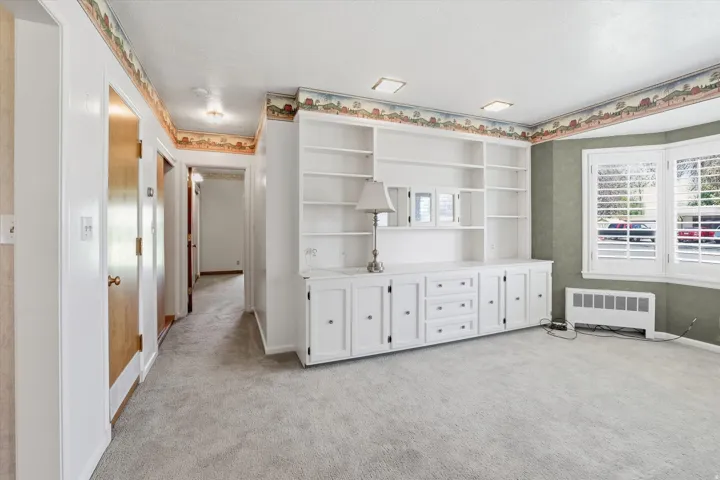 Integrated white cabinetry with shelving, a bay window featuring white plantation shutters, light gray carpet flooring, recessed ceiling lighting, and a corridor with natural wood doors