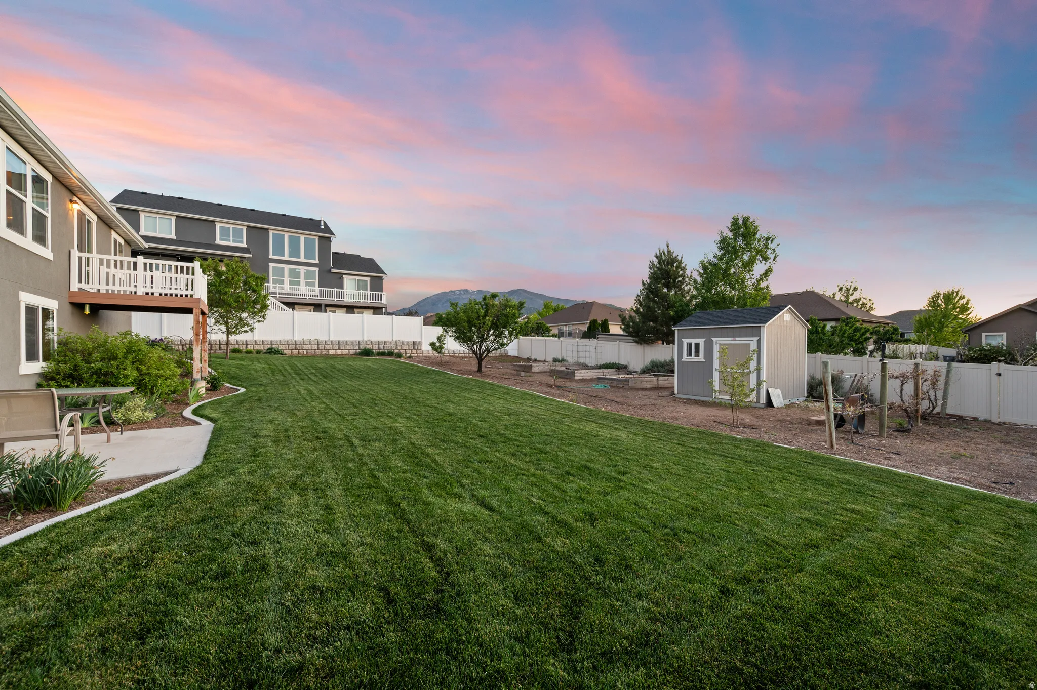 Expansive backyard featuring a manicured lawn, a concrete patio, a raised deck with white railings, a detached storage shed, and a white privacy fence