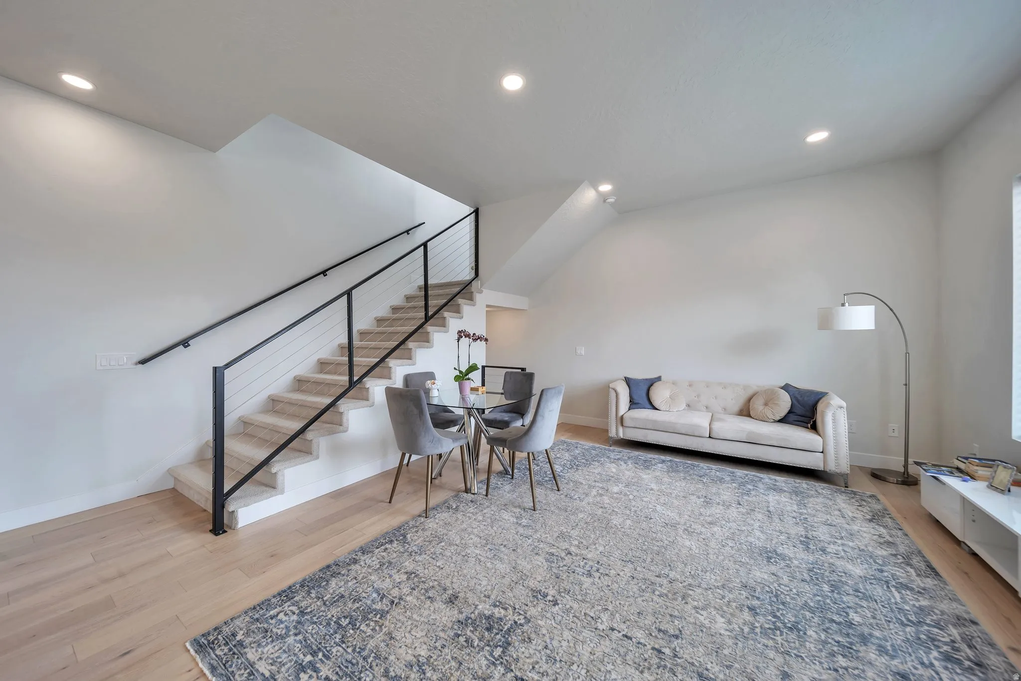Open-concept living area with wood-finish flooring, recessed lighting, and a modern staircase featuring a black metal railing with cable infill