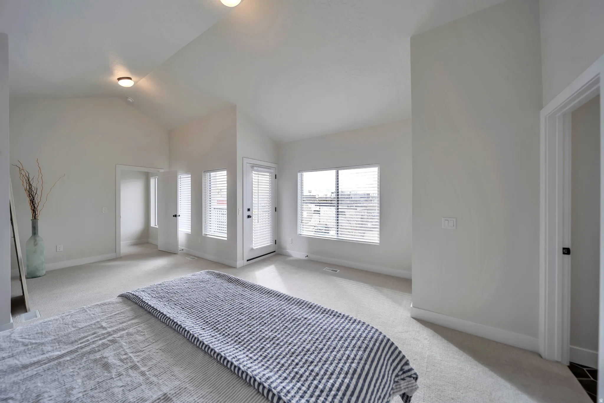 Spacious room featuring vaulted ceilings, light-colored carpeting, multiple windows with blinds, a glass-paned exterior door, and an interior doorway with a white frame
