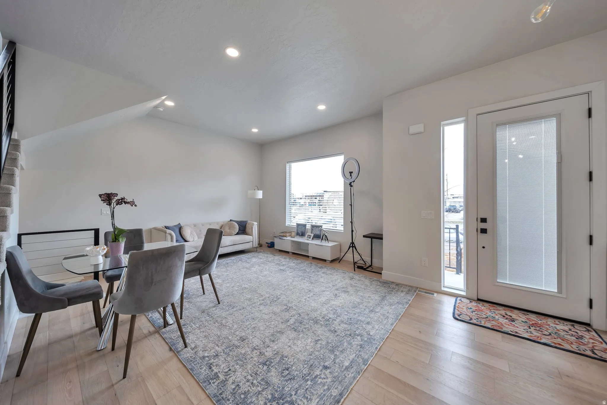 Bright open-concept living area featuring light wood-finish flooring, recessed lighting, and a large front window with blinds