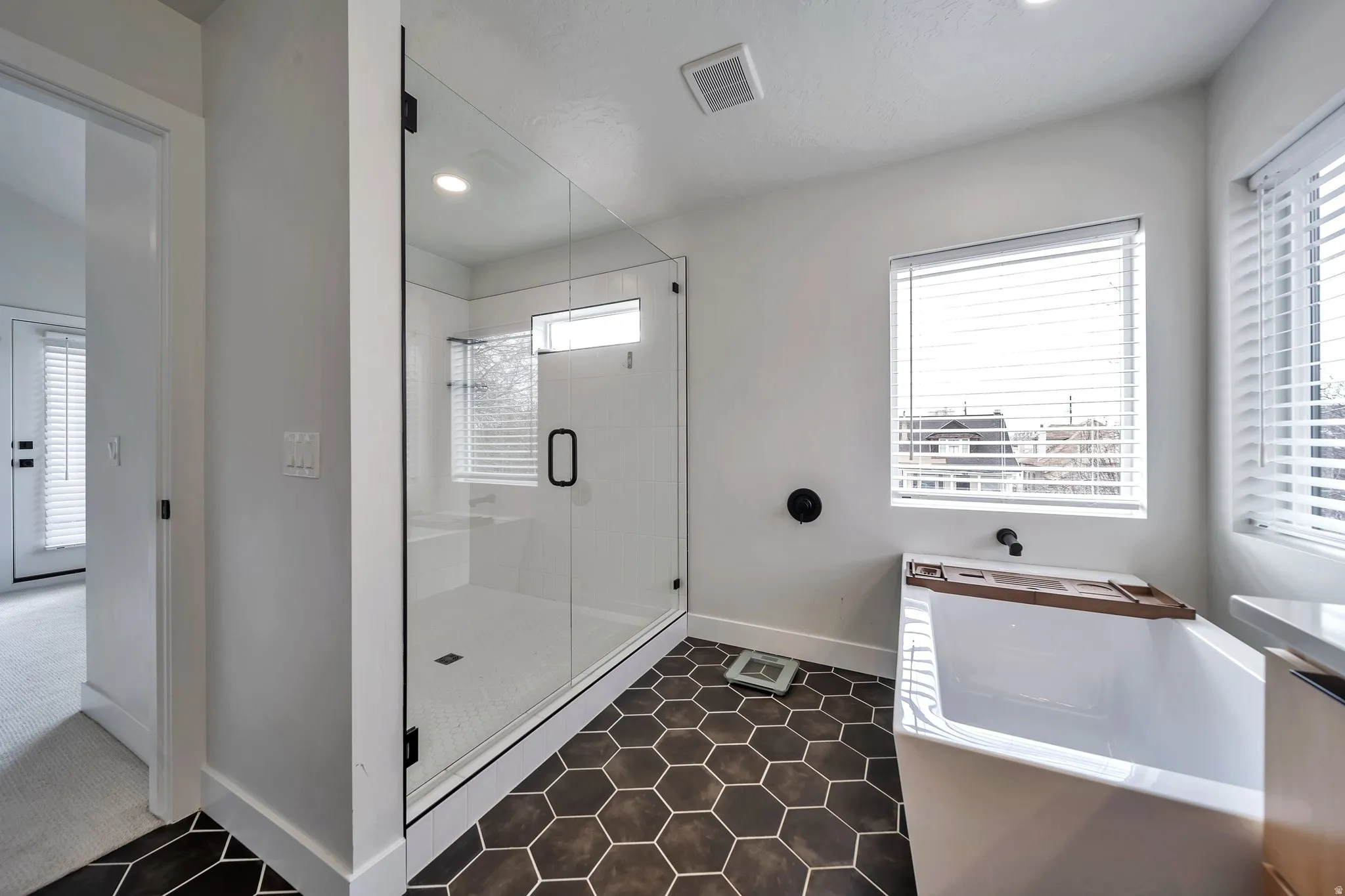Modern bathroom featuring dark hexagon floor tiling, a clear glass shower enclosure with white subway tile, a freestanding soaking tub, and dual windows with blinds
