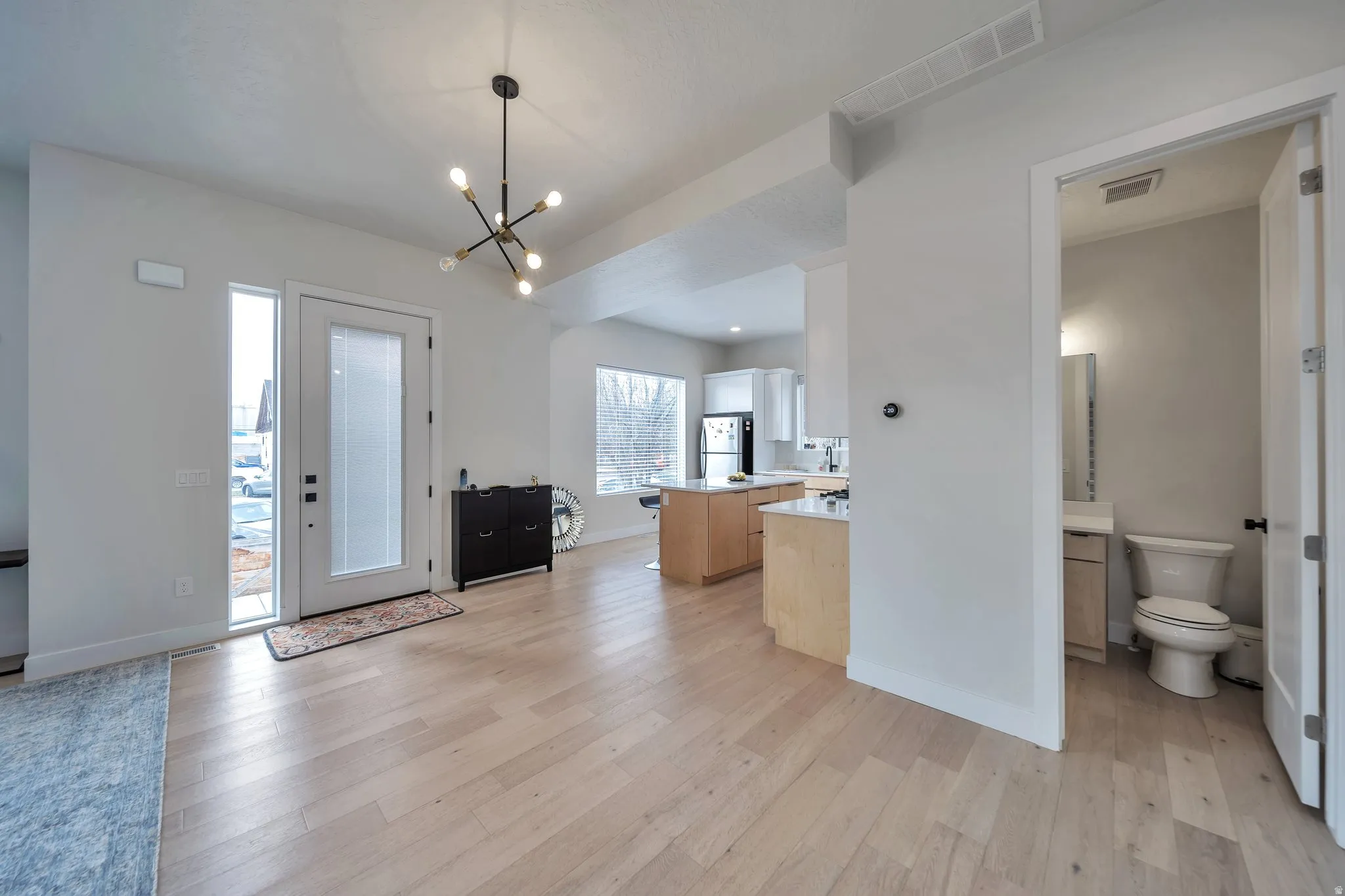 Spacious open-concept living area featuring light wood-finish flooring, a modern chandelier, and a bright entryway with a full-light door and vertical sidelight