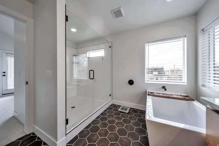 Modern bathroom featuring dark hexagon floor tiling, a clear glass shower enclosure with white subway tile, a freestanding soaking tub, and dual windows with blinds
