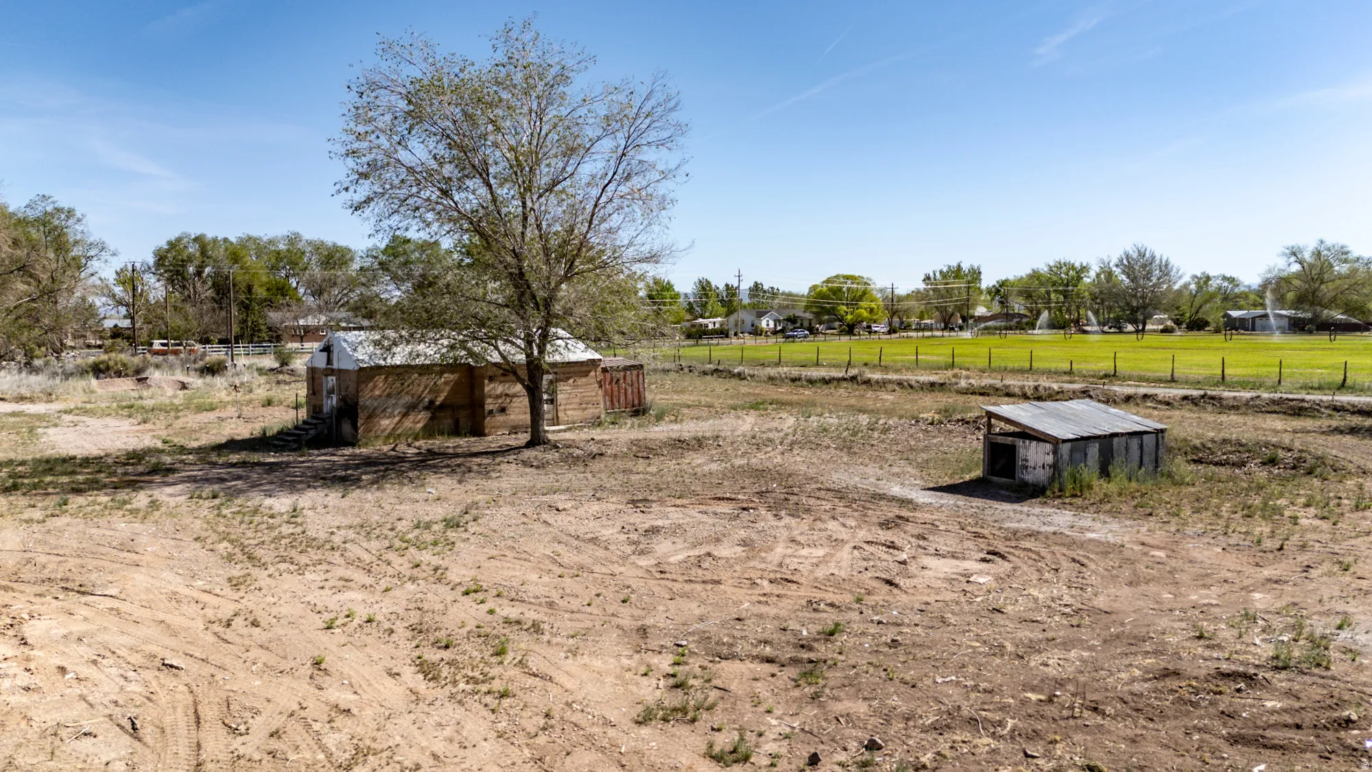Expansive dirt lot featuring a mature tree, two outbuildings, and a perimeter fence bordering a grassy field