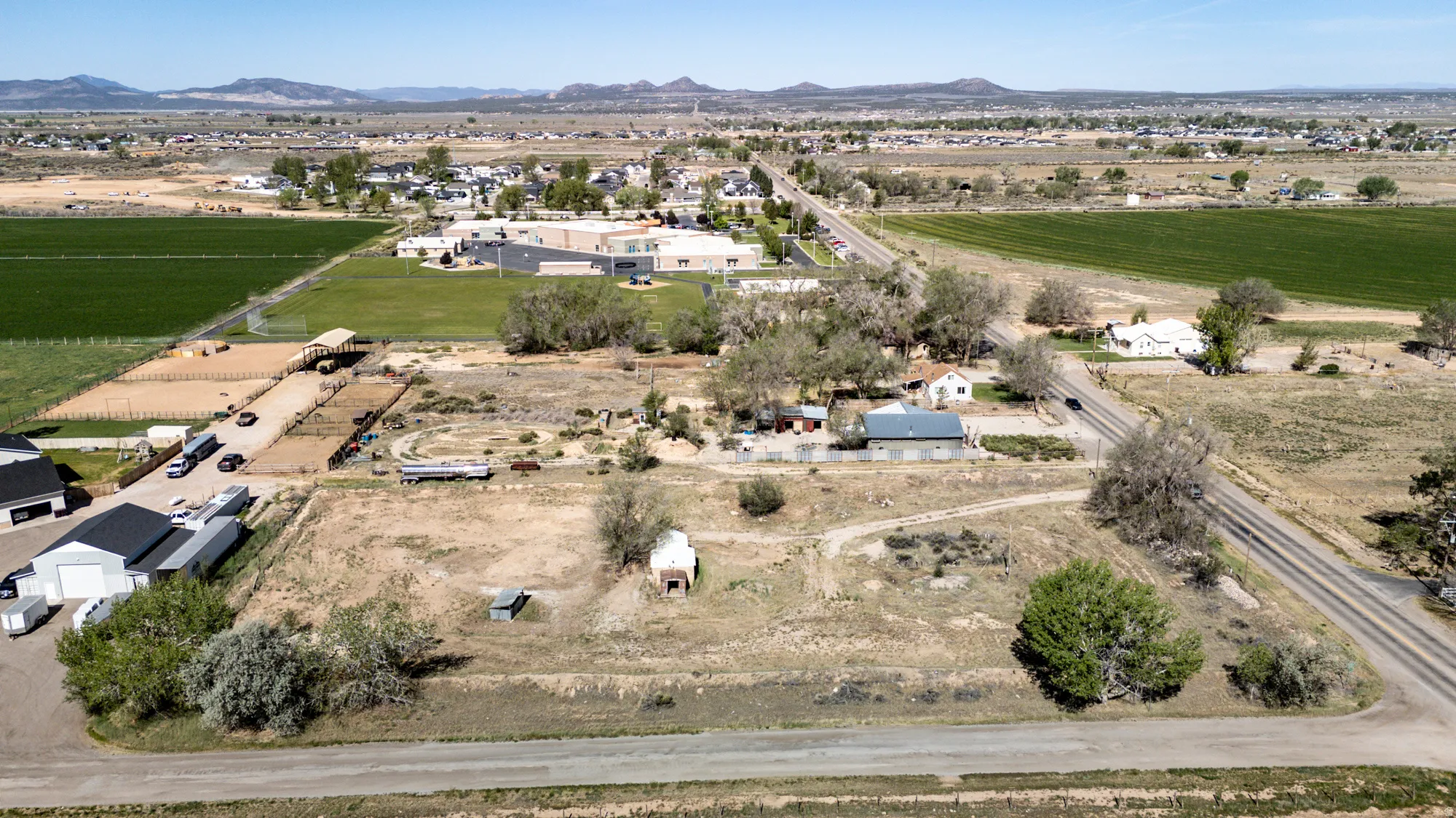 Expansive agricultural land featuring a verdant field, multiple outbuildings with dark roofs, and a large unpaved area