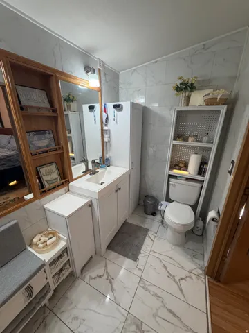 Bathroom featuring white marble-look floor and wall tiles, a white vanity with integrated sink, and a mirrored wall cabinet with wood shelving