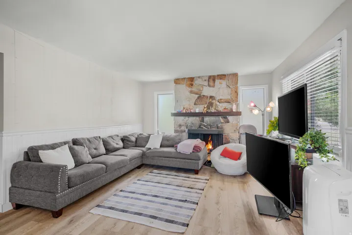 Living area featuring a stone fireplace with a wood mantle, wood-finish flooring, white beadboard wainscoting, and a large window with horizontal blinds